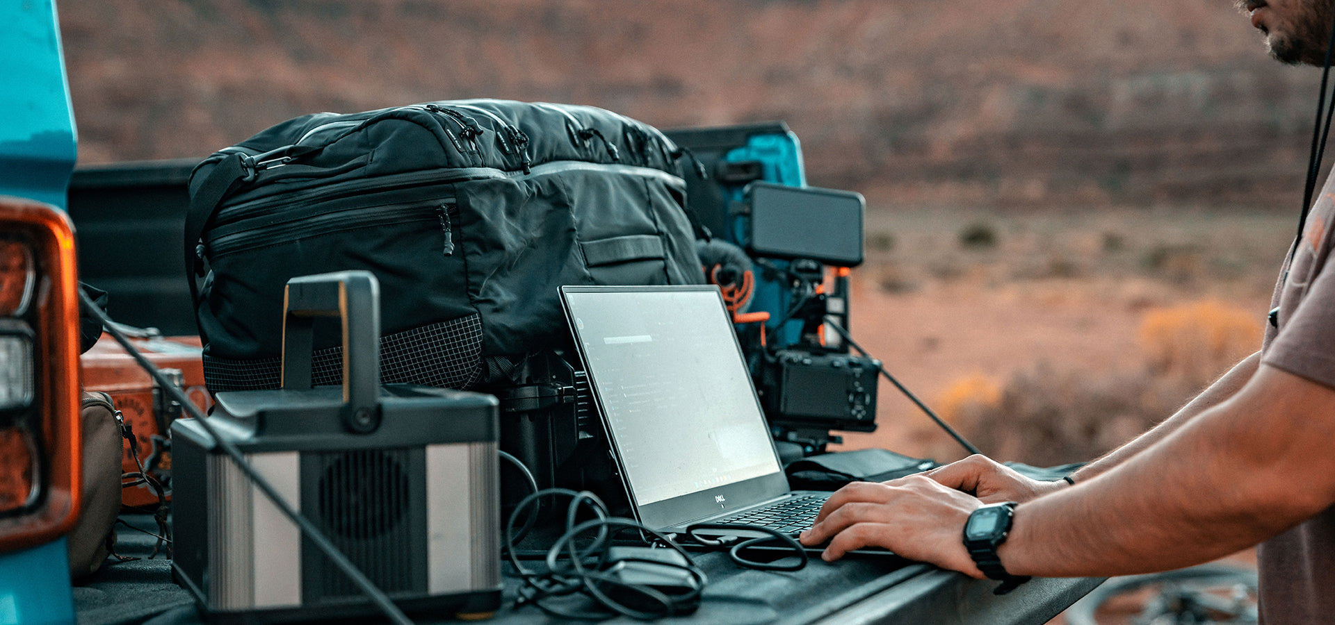 Man in desert setting working on laptop from back of his truck, filled with bags and equipment